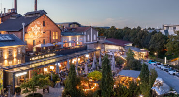 Ein Brauerei- und Restaurantkomplex im Freien mit Backsteingebäuden, großen Leuchtreklamen, Bäumen, Lichterketten und Menschen, die in der Abenddämmerung auf den Terrassen speisen. Auf der rechten Seite sind Autos geparkt, und der Platz ist von Grün umgeben.