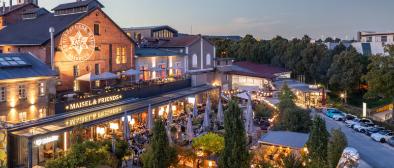 Ein Brauerei- und Restaurantkomplex im Freien mit Backsteingebäuden, großen Leuchtreklamen, Bäumen, Lichterketten und Menschen, die in der Abenddämmerung auf den Terrassen speisen. Auf der rechten Seite sind Autos geparkt, und der Platz ist von Grün umgeben.