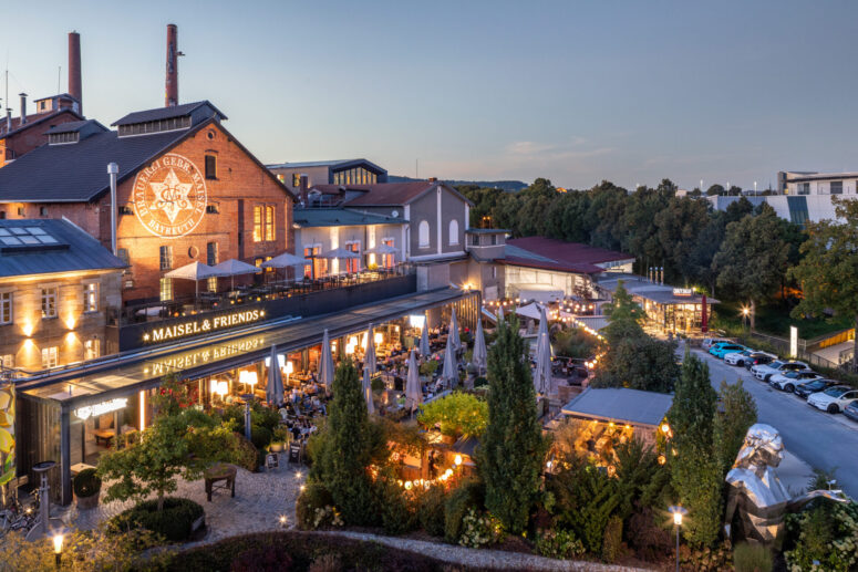 Ein Brauerei- und Restaurantkomplex im Freien mit Backsteingebäuden, großen Leuchtreklamen, Bäumen, Lichterketten und Menschen, die in der Abenddämmerung auf den Terrassen speisen. Auf der rechten Seite sind Autos geparkt, und der Platz ist von Grün umgeben.