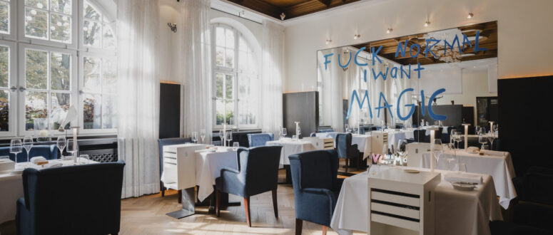 Ein stilvolles Restaurant mit weißen Tischtüchern, blauen Stühlen, großen Bogenfenstern und einer Holzdecke. Auf einem Spiegel an der Wand steht mit blauer Farbe die Aufschrift FUCK NORMAL I WANT MAGIC.