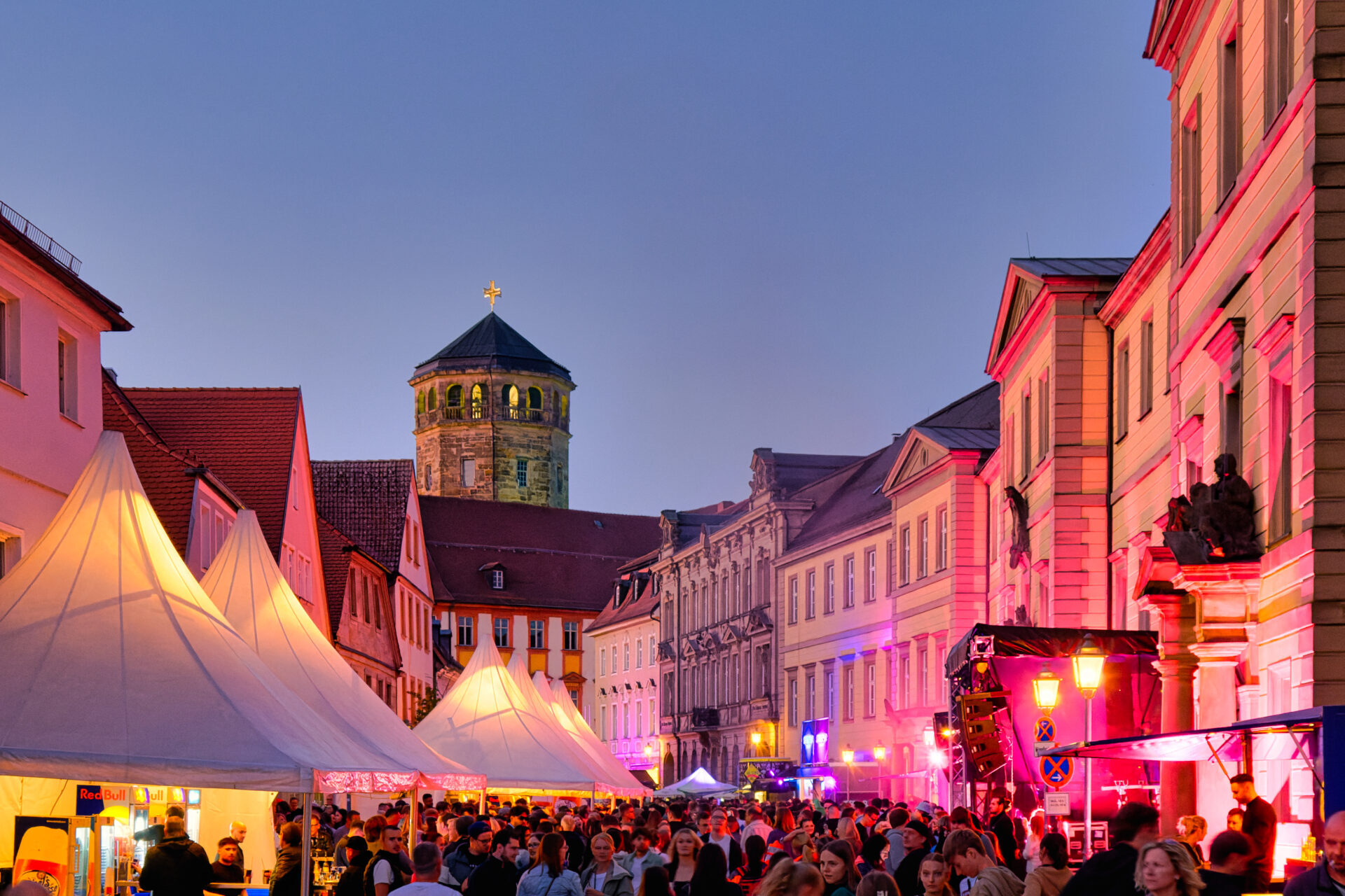 Ein lebendiges Bayreuther Straßenfest in der Abenddämmerung mit Menschenmassen, weißen Zelten, bunten Lichtern, die historische Gebäude beleuchten, und einem steinernen Kirchturm im Hintergrund.
