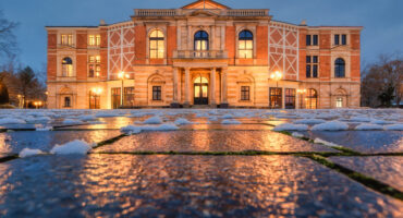 Ein großes, beleuchtetes Festspielhaus mit klassischer Architektur spiegelt sich in der Abenddämmerung auf nassen, schneebedeckten Pflastersteinen. Der Himmel ist bewölkt und blau, und die Fenster des Gebäudes leuchten warm.
