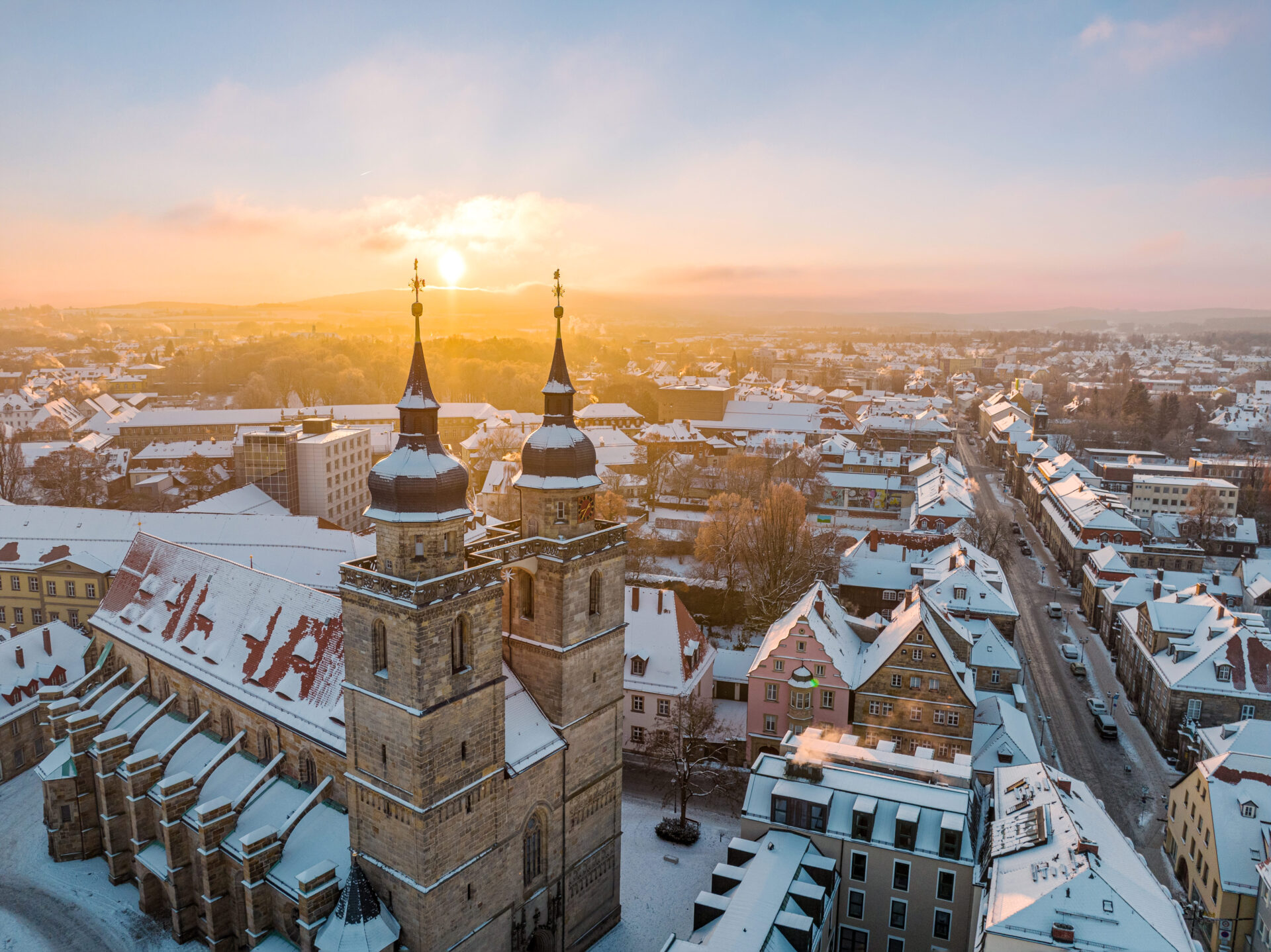 Luftaufnahme einer verschneiten Stadt bei Sonnenaufgang, mit einer großen Kirche mit zwei Türmen im Vordergrund - perfekt für einen Kurzurlaub in Bayreuth - und Häusern und Straßen, die sich unter einem pastellfarbenen Himmel in die Ferne erstrecken.