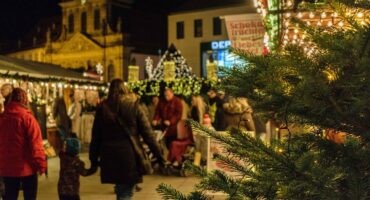 Bild vom Christkindlesmarkt Bayreuth durch einen Weihnachtsbaum fotografiert. Zu sehen sind über den schön beleuchteten Markt schlendernde Menschen und die Kirche im Hintergrund.