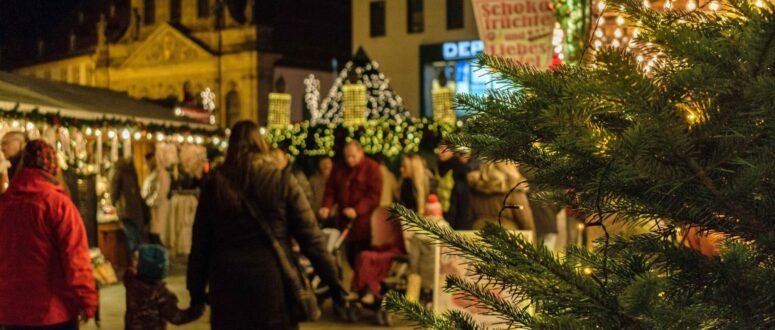 Bild vom Christkindlesmarkt Bayreuth durch einen Weihnachtsbaum fotografiert. Zu sehen sind über den schön beleuchteten Markt schlendernde Menschen und die Kirche im Hintergrund.