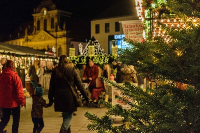 Bild vom Christkindlesmarkt Bayreuth durch einen Weihnachtsbaum fotografiert. Zu sehen sind über den schön beleuchteten Markt schlendernde Menschen und die Kirche im Hintergrund.