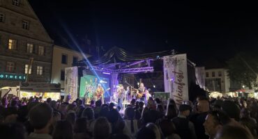 Eine große Menschenmenge schaut einer Live-Band zu, die während des Weinfestes auf einer Freilichtbühne auftritt, während im Hintergrund Gebäude, Lichter und Festivalbanner zu sehen sind.