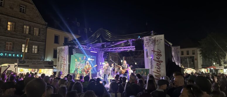 Eine große Menschenmenge schaut einer Live-Band zu, die während des Weinfestes auf einer Freilichtbühne auftritt, während im Hintergrund Gebäude, Lichter und Festivalbanner zu sehen sind.