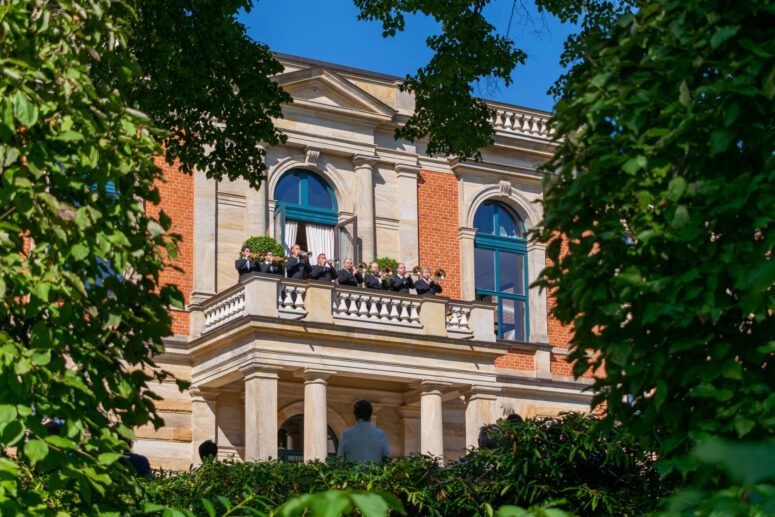 Eine Gruppe von Menschen steht an einem sonnigen Tag auf dem großen steinernen Balkon des historischen Bayreuther Festspielhauses von Richard Wagner, mit gewölbten Fenstern und grünen Bäumen um sie herum.