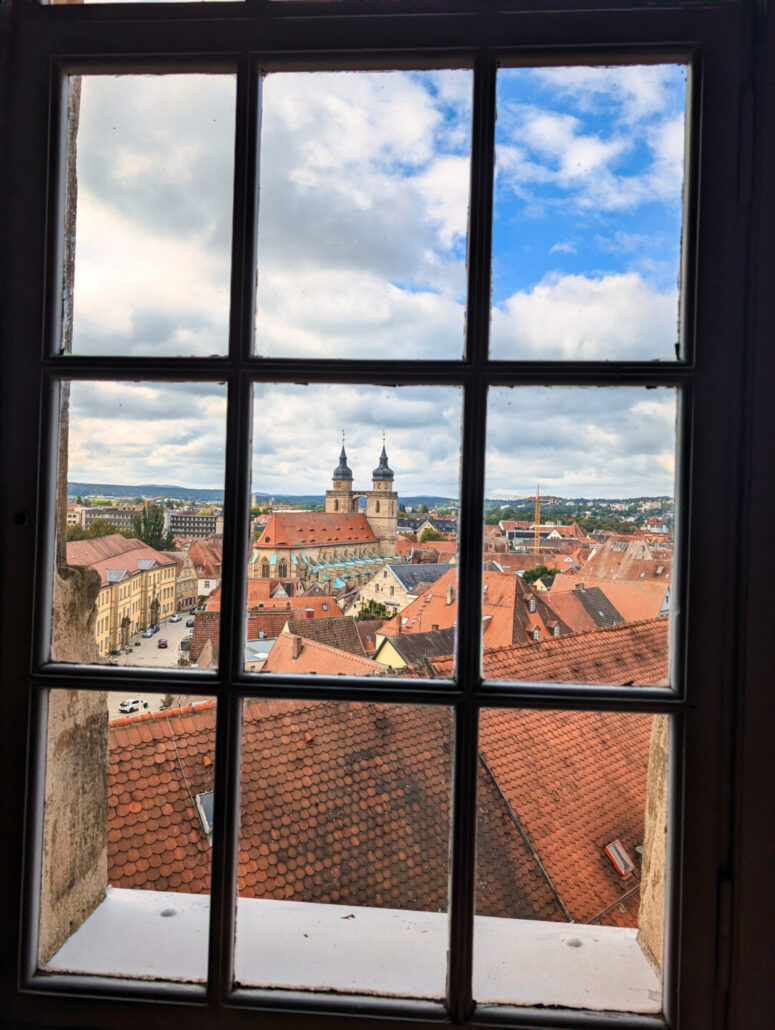 Blick durch ein Fenster mit Scheiben auf eine europäische Stadt mit roten Ziegeldächern, historischen Gebäuden und einer Kirche mit zwei Türmen unter einem teilweise bewölkten Himmel.