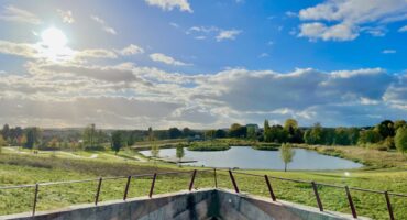Blick auf eine grasbewachsene Landschaft mit einem Teich, Bäumen und blauem Himmel mit Wolken, gesehen hinter einem Betongeländer in der hellen Nachmittagssonne - perfekt für schöne Winterspaziergänge.