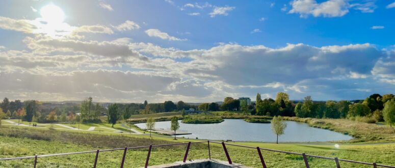 Blick auf eine grasbewachsene Landschaft mit einem Teich, Bäumen und blauem Himmel mit Wolken, gesehen hinter einem Betongeländer in der hellen Nachmittagssonne - perfekt für schöne Winterspaziergänge.