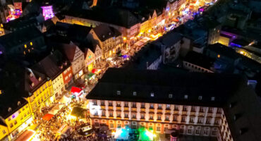 Ein lebendiges Luftbild von Bayreuth bei Nacht zeigt hell erleuchtete Straßen, historische Gebäude und eine große Menschenmenge, die sich auf einem beleuchteten Platz in der Nähe des berühmten Bayreuther Ladens zu einer lebhaften Freiluftveranstaltung oder einem Festival versammelt hat.