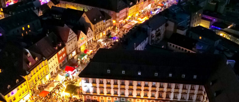 Ein lebendiges Luftbild von Bayreuth bei Nacht zeigt hell erleuchtete Straßen, historische Gebäude und eine große Menschenmenge, die sich auf einem beleuchteten Platz in der Nähe des berühmten Bayreuther Ladens zu einer lebhaften Freiluftveranstaltung oder einem Festival versammelt hat.