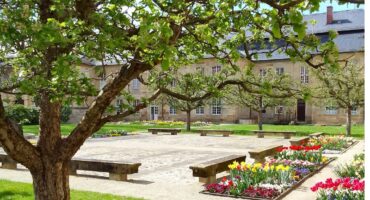 Ein friedlicher Garten mit blühenden Blumenbeeten, Steinbänken und belaubten Bäumen heißt den Frühling willkommen. Im Hintergrund steht ein historisches Steingebäude mit vielen Fenstern und einem Schieferdach unter einem strahlenden Himmel.
