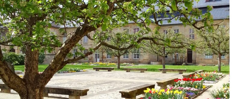 Ein friedlicher Garten mit blühenden Blumenbeeten, Steinbänken und belaubten Bäumen heißt den Frühling willkommen. Im Hintergrund steht ein historisches Steingebäude mit vielen Fenstern und einem Schieferdach unter einem strahlenden Himmel.