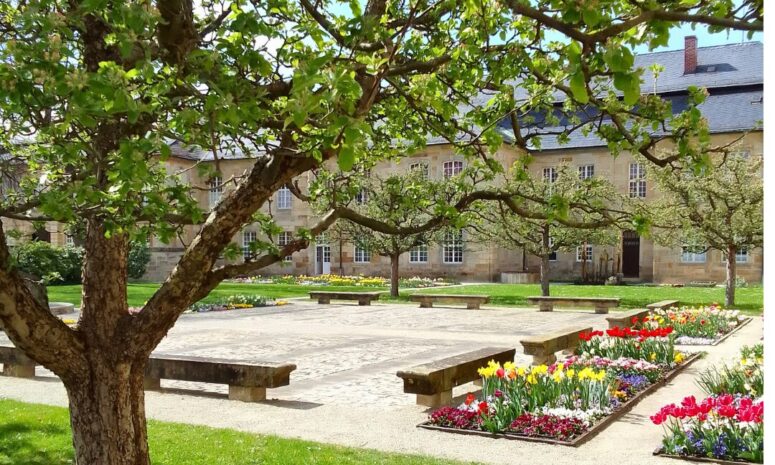 Ein friedlicher Garten mit blühenden Blumenbeeten, Steinbänken und belaubten Bäumen heißt den Frühling willkommen. Im Hintergrund steht ein historisches Steingebäude mit vielen Fenstern und einem Schieferdach unter einem strahlenden Himmel.