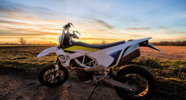 Ein weißes Motorrad steht auf einem unbefestigten Weg mit einem Feld und Bäumen im Hintergrund, beleuchtet vom warmen Licht eines farbenfrohen Sonnenuntergangs.