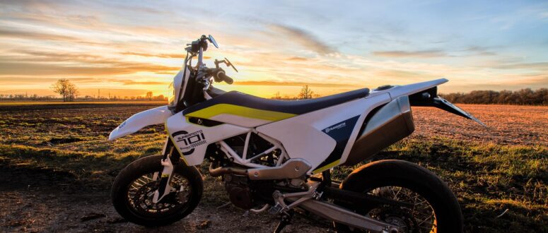 Ein weißes Motorrad steht auf einem unbefestigten Weg mit einem Feld und Bäumen im Hintergrund, beleuchtet vom warmen Licht eines farbenfrohen Sonnenuntergangs.