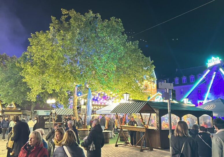 Ein lebendiges Fest unter freiem Himmel bei Nacht, wie das Bayreuther Volksfest der Genussgärten, mit Menschenmassen, beleuchteten Bäumen, bunten Lichtern, Verkaufsständen und Gebäuden im Hintergrund - eine lebendige und festliche Szene unter dem Nachthimmel.
