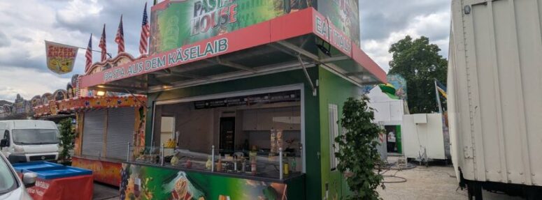 Ein bunter Imbissstand mit grüner und roter Beschriftung steht geschlossen auf dem Festplatz der Genussgärten Bayreuther Volksfest. In der Nähe befinden sich weitere Stände, ein weißer Lastwagen und einige Bäume unter einem bewölkten Himmel.