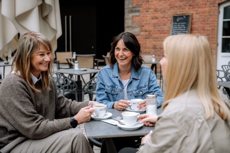 Drei Frauen sitzen an einem Cafétisch im Freien, lächeln und unterhalten sich mit einer Tasse Kaffee in der Hand. Sie wirken entspannt und glücklich, mit einer Backsteinmauer und Café-Stühlen im Hintergrund.