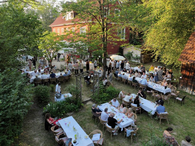 Eine Versammlung im Freien mit vielen Menschen, die an weiß gedeckten Tischen in einem Garten sitzen, umgeben von Bäumen und Grün, mit einem großen roten Gebäude im Hintergrund.
