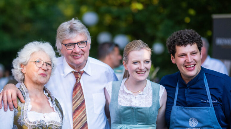 Vier Erwachsene, drei von ihnen in traditioneller bayerischer Kleidung und einer in der Uniform eines Kochs, stehen lächelnd und mit umschlungenen Armen im Freien zusammen. Der Hintergrund ist üppig und grün.