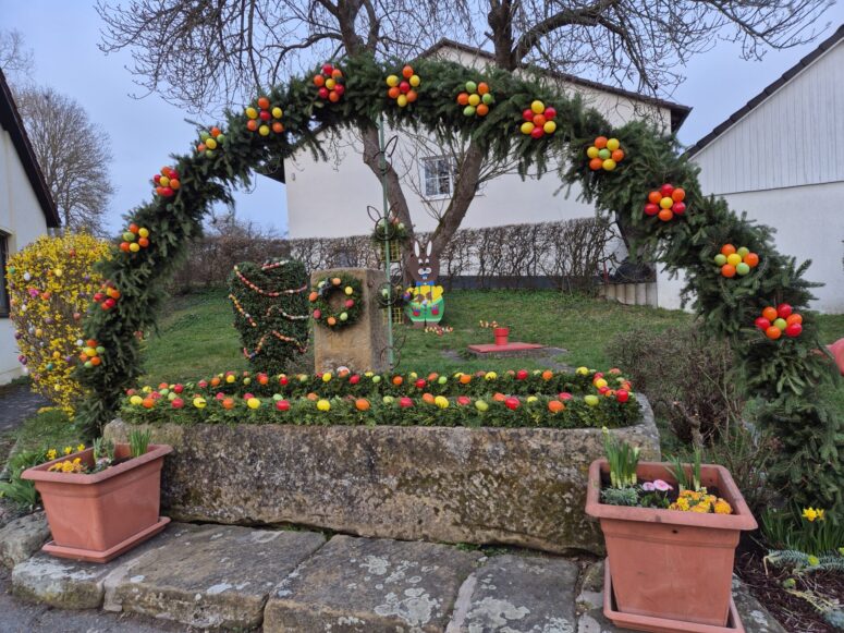 Ein festlicher Garten zeigt einen großen Osterbrunnenbogen, der mit grünem Laub und bunten Ostereiern geschmückt ist und von Blumenkübeln flankiert wird. Im Hintergrund sind gestutzte Sträucher, ein Holzhase und Häuser zu sehen.
