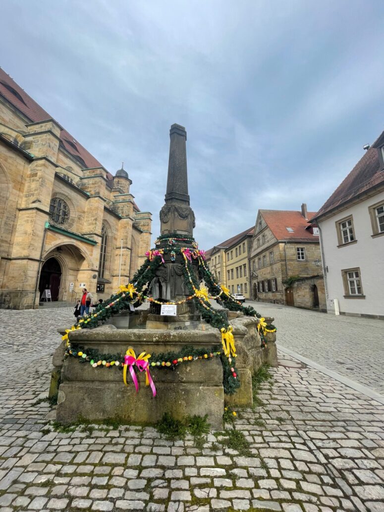 Ein mit Girlanden, bunten Bändern und Ostereiern geschmückter Steinbrunnen steht auf einem kopfsteingepflasterten Platz, umgeben von historischen Gebäuden, unter einem bewölkten Himmel.
