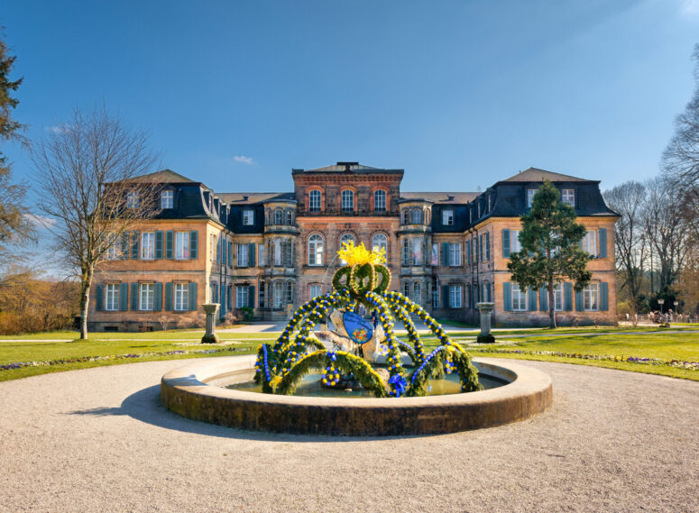 Hinter einem runden, mit leuchtend gelben und blauen Blumen geschmückten Springbrunnen, umgeben von einem gepflegten Rasen und Bäumen, steht ein prächtiges historisches Herrenhaus mit vielen Fenstern unter einem klaren blauen Himmel.