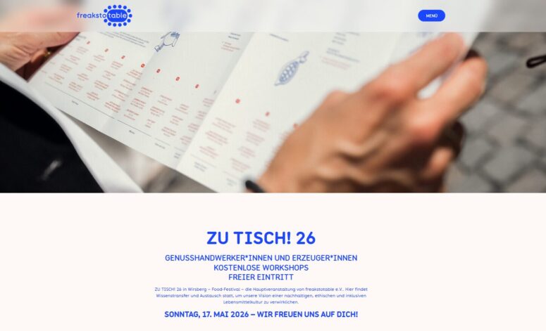 Nahaufnahme einer Person, die ein gefaltetes Veranstaltungsprogramm oder eine Speisekarte in der Hand hält, darunter Text auf einer Website in deutscher Sprache über "Zu Tisch! 26", eine Veranstaltung mit Workshops und freiem Eintritt am 17. Mai 2026.
