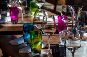 Nahaufnahme eines Restauranttisches mit bunten Glasbechern, Weingläsern, einer Glaskaraffe und ordentlich aufgerollten weißen Servietten auf einer Holzfläche. Das Sonnenlicht hebt die leuchtenden Farben und Glasreflexe hervor.