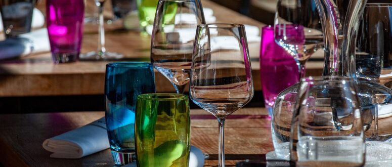 Nahaufnahme eines Restauranttisches mit bunten Glasbechern, Weingläsern, einer Glaskaraffe und ordentlich aufgerollten weißen Servietten auf einer Holzfläche. Das Sonnenlicht hebt die leuchtenden Farben und Glasreflexe hervor.