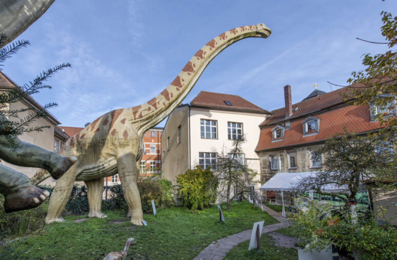 Ein großes, lebensgroßes Dinosauriermodell steht in einem grasbewachsenen Innenhof, umgeben von historischen Gebäuden mit roten Dächern unter einem strahlend blauen Himmel.