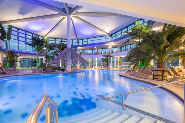 Geräumiger Innenpool mit türkisfarbenem Wasser, umgeben von Liegestühlen, Palmen und großen Fenstern. Die moderne Architektur mit einer gewölbten Decke und sanfter violetter Beleuchtung schafft eine entspannende, tropische Atmosphäre.