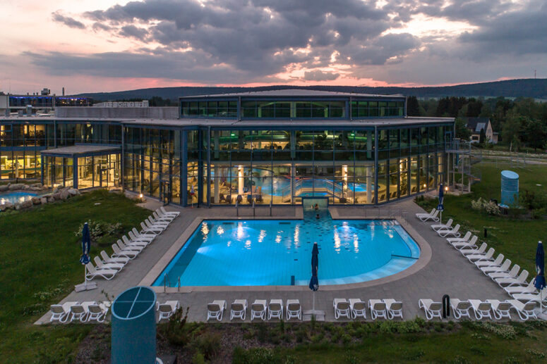 Ein modernes Glasgebäude mit großen Fenstern blickt auf einen Außenpool, der bei Sonnenuntergang von weißen Liegestühlen umgeben ist, mit ein paar Sonnenschirmen und grünem Rasen im Hintergrund.