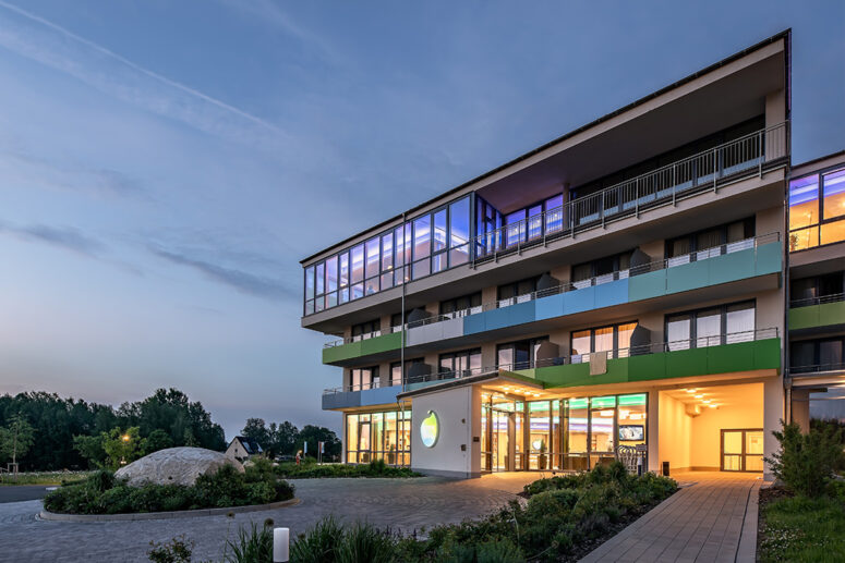 Ein modernes, mehrstöckiges Hotel mit großen Glasfenstern und farbenfrohen Balkonen wird in der Abenddämmerung beleuchtet, davor befinden sich angelegte Gärten und eine kreisförmige Auffahrt. Im Hintergrund sind Bäume und ein klarer Himmel zu sehen.