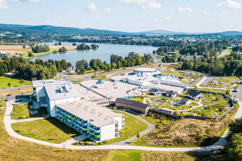 Luftaufnahme eines großen Ferienkomplexes mit mehreren Gebäuden, Außenpools und Grünanlagen in der Nähe eines Sees, umgeben von grünen Feldern, Bäumen und sanften Hügeln unter einem blauen Himmel mit vereinzelten Wolken.