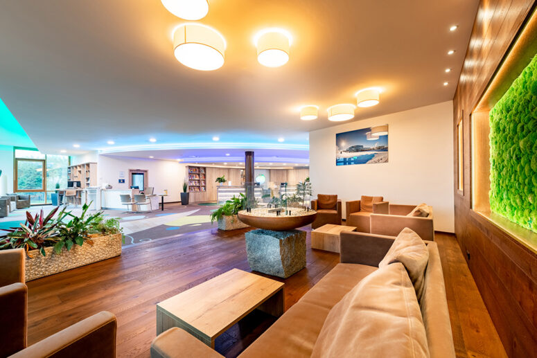 Eine moderne, geräumige Lobby mit beigen Sofas, Holzfußboden, Pflanzen und einer grünen Mooswand. Warme Deckenleuchten und natürliches Licht erhellen den Raum, mit Bücherregalen und einem Empfangstresen im Hintergrund.