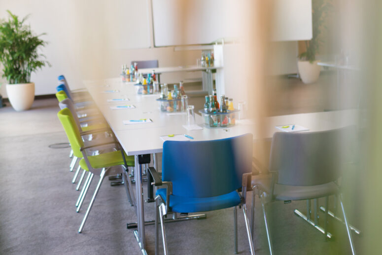 Ein heller Konferenzraum mit zwei langen, U-förmig angeordneten Tischen, umgeben von bunten Stühlen. Auf den Tischen befinden sich Notizblöcke, Stifte und Getränke. Im Hintergrund stehen Topfpflanzen.