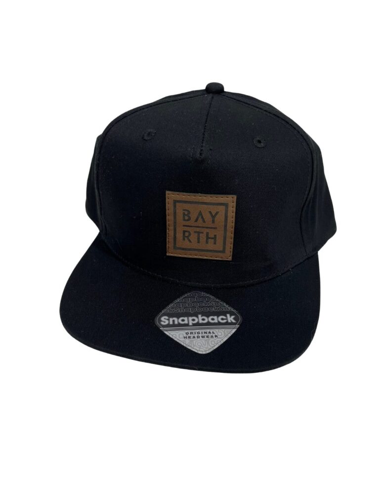 Eine schwarze Snapback-Kappe mit flacher Krempe und einem braunen Lederaufnäher auf der Vorderseite mit dem Text BAY RTH-erhältlich ab sofort im Bayreuth Shop. An der Krempe ist ein silberner Snapback-Aufkleber angebracht.