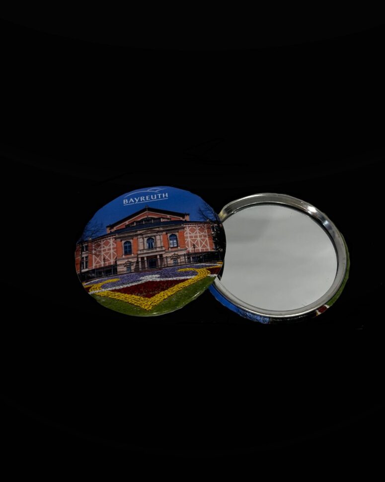 Ein runder Kompaktspiegel aus dem Bayreuth-Shop wird aufgeklappt und enthüllt seine spiegelnde Seite. Das Cover zeigt ein Foto des Bayreuther Festspielhauses und ein buntes Blumenbeet vor schwarzem Hintergrund.