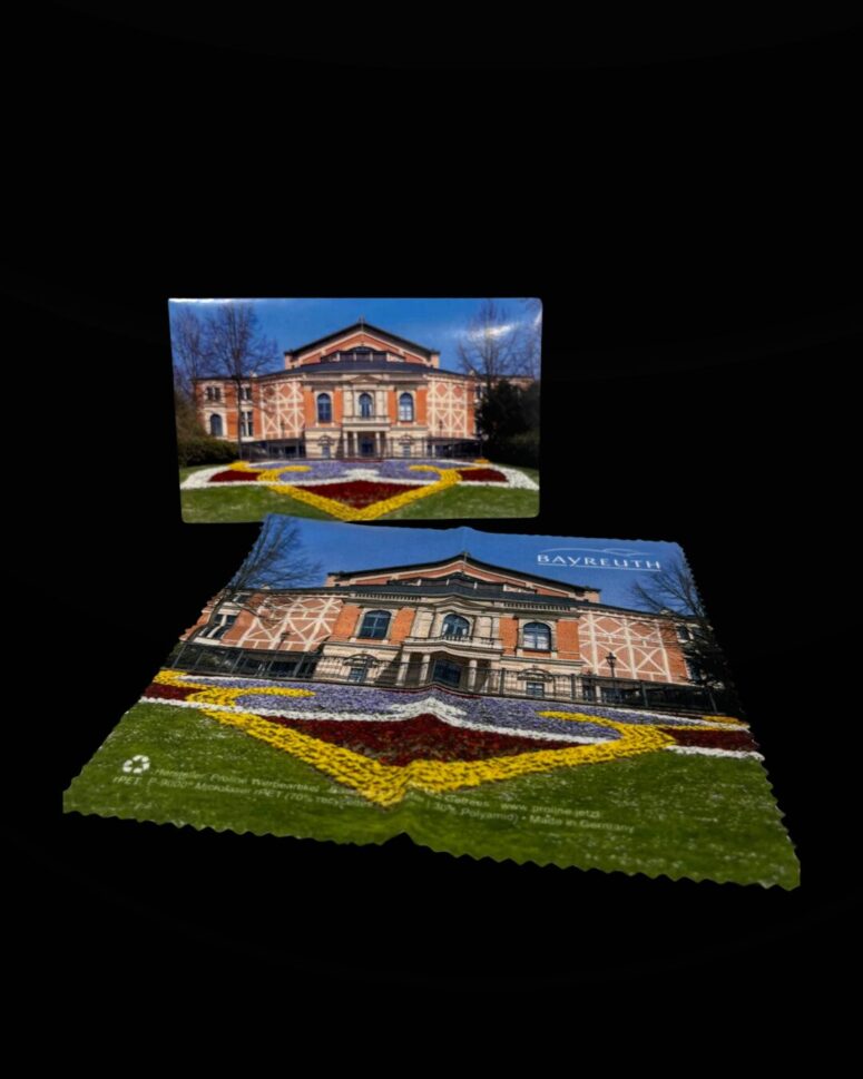 Ein gedrucktes Foto und ein Souvenirtuch aus dem Bayreuth-Shop, die beide ein Gebäude mit einer gemusterten Fassade und bunten Blumenbeeten davor zeigen, sind vor einem schwarzen Hintergrund zu sehen.