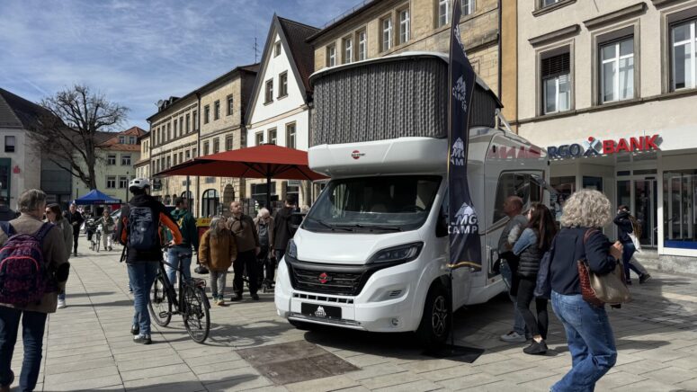 Eine Gruppe von Menschen versammelt sich während der Mobilitätstage um ein großes weißes Wohnmobil, das auf einer Straße in der Stadt vor Geschäften und historischen Gebäuden geparkt ist; einige gehen, unterhalten sich oder stehen mit Fahrrädern.
