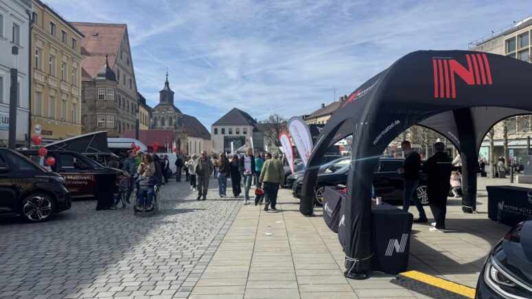 Auf einem mit Kopfsteinpflaster gepflasterten Stadtplatz findet eine lebhafte Autoausstellung der Mobilitätstage statt, bei der Menschen zwischen Ausstellungszelten, Autos und roten Luftballons spazieren gehen, mit historischen Gebäuden und einer Kirche im Hintergrund unter einem teilweise bewölkten Himmel.