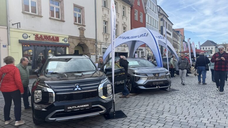 Bei den Mobilitätstagen, einer Freiluftveranstaltung auf einem Stadtplatz mit bunten Gebäuden und Geschäften im Hintergrund, gehen die Besucher an zwei Mitsubishi-Fahrzeugen vorbei, die unter Zelten ausgestellt sind.