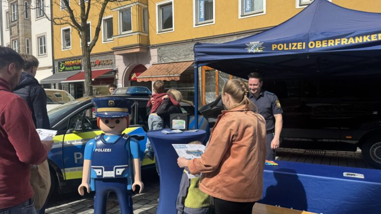 Während der Mobilitätstage interagieren die Besucher an einem Informationsstand der Polizei im Freien mit einem blauen Zelt mit der Aufschrift POLIZEI OBERFRANKEN. Eine große Playmobil-Polizeifigur und ein Polizeiauto sind zu sehen, während ein Polizist mit einem Besucher spricht.