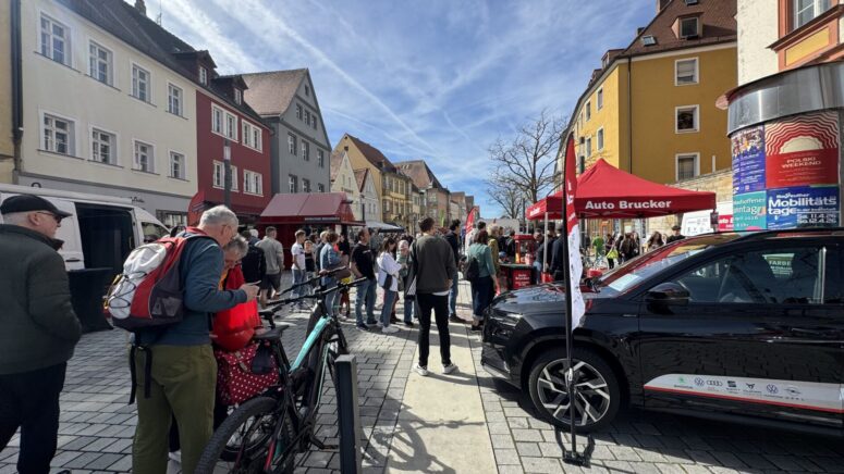 Während der Mobilitätstage findet ein lebhaftes Straßenfest statt, bei dem sich Menschen um Stände und Auslagen scharen, darunter ein rotes Zelt und ein geparktes schwarzes Auto. Bunte Gebäude und ein strahlend blauer Himmel bilden die Kulisse, während im Vordergrund Fahrräder geparkt sind.