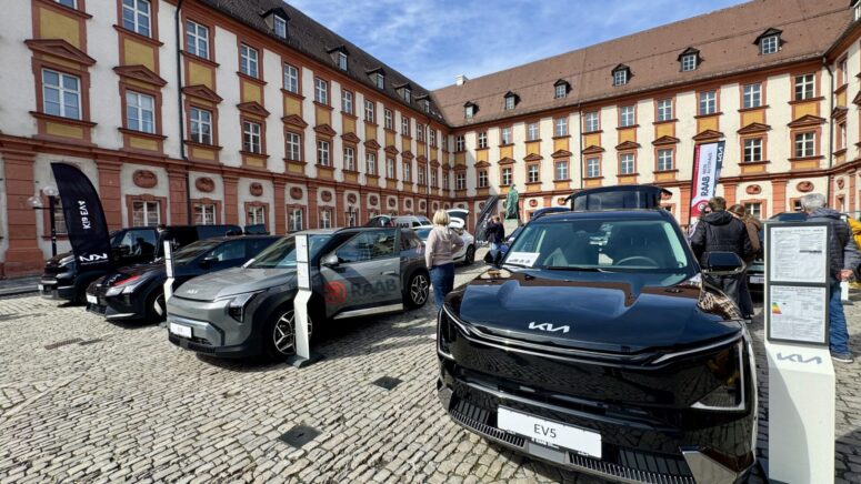 Mehrere Kia-Elektrofahrzeuge sind während der Mobilitätstage in einem historischen Hof mit Kopfsteinpflaster ausgestellt, der von einem großen rot-beigen Gebäude umgeben ist. Die Menschen gehen umher und begutachten die Autos unter einem sonnigen Himmel.
